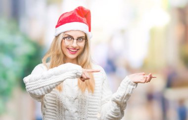 Hayret ve el ile sunulması ve parmak ile işaret ederken kameraya gülümseyen izole arka plan üzerinde Noel şapka giyen genç güzel sarışın kadın.