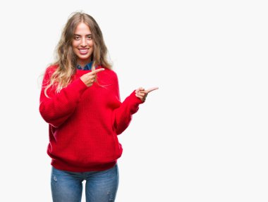 Gülümseyen ve iki elleri ve parmakları ile tarafına işaret kamera bakarak izole arka plan üzerinde kış kazak giyen güzel genç sarışın kadın.