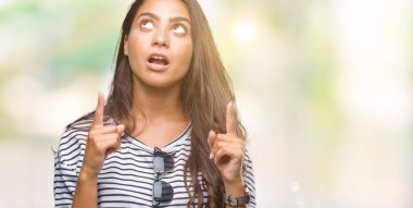 Genç güzel Arap kadın hayrete ve yukarıya bakıyor ve parmakları ile işaret ve silah kaldırdı sürpriz izole arka plan üzerinde güneş gözlüğü takıyor.