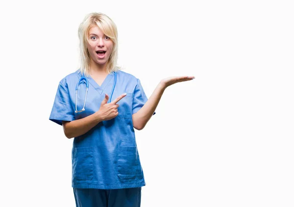 驚くし 手とを提示 指で指している間カメラに笑顔孤立の背景に医療制服を着て美しい金髪女医女性 — ストック写真