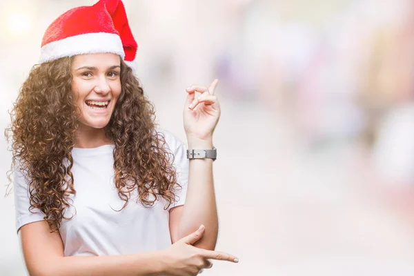 若いブルネットの少女の顔に大きな笑みを浮かべて隔離された背景にクリスマスの帽子を着てカメラを見て側の手と指で指す — ストック写真