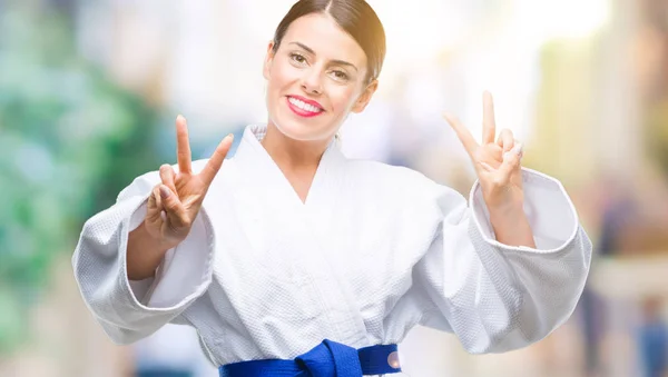 Young beautiful woman wearing karate kimono uniform over isolated background smiling looking to the camera showing fingers doing victory sign. Number two.