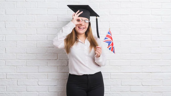 指の間から見て目に手で サインをして笑っている幸せそうな顔でイギリスの旗を保持している大学院のキャップを身に着けている白いレンガ壁を越えて若い成人女性 — ストック写真