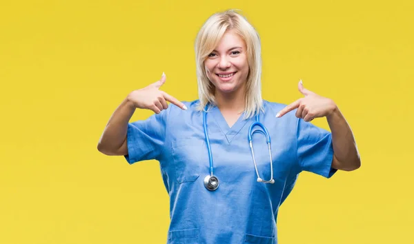 美しい金髪女医女性の顔に笑顔と自信を持って 誇りと幸せな指で自分を指している孤立した背景に医療の制服を着て — ストック写真