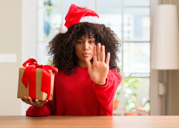 African american woman holding present wearing christmas red hat with open hand doing stop sign with serious and confident expression, defense gesture