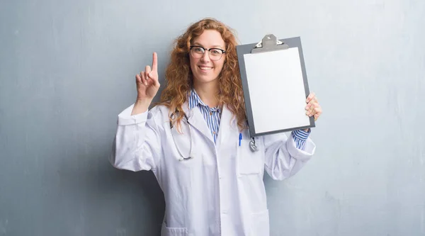 Joven Doctora Pelirroja Sobre Pared Grunge Gris Mostrando Portapapeles Sorprendida —  Fotos de Stock