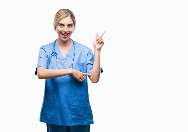 美しい金髪女医外科医看護師女性の顔に大きな笑みを浮かべて隔離された背景にカメラを見ている側の手と指で指す — ストック写真