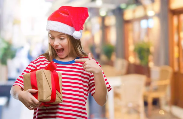 非常に幸せな手と指で指している孤立した背景の上クリスマス帽子 持株ギフトを着た美しい少女 — ストック写真