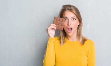 Grunge güzel genç kadın çikolatayı bir sürpriz yüz, korkuyor ve heyecanlı korku ifadesi ile şok korkmuş yeme duvar gri