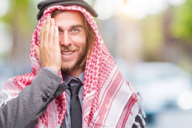 Genç yakışıklı Arap adam uzun saçlı bir göz ile yüz ve sürpriz duygu kendinden emin gülümseme ile el kapsayan izole arka plan üzerinde keffiyeh giyiyor.