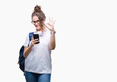 Sırt çantası, kulaklık ve akıllı telefon parmak, mükemmel sembolü ile ok işareti yapıyor izole arka plan üzerinde giyen genç esmer öğrenci kız