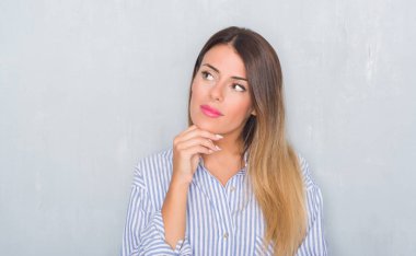 Young adult woman over grunge grey wall wearing business shirt with hand on chin thinking about question, pensive expression. Smiling with thoughtful face. Doubt concept. clipart