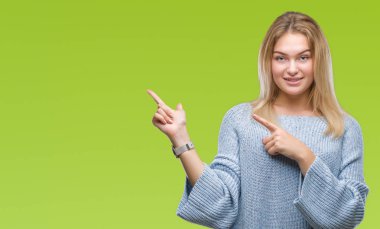 Gülümseyen ve iki elleri ve parmakları ile tarafına işaret kamera bakarak izole arka plan üzerinde kış kazak giyen genç beyaz kadın.