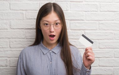 Kredi kartı bir sürpriz yüz, korkuyor ve heyecanlı korku ifadesi ile şok korkmuş tutan tuğla duvarın üzerinden Genç Çinli kadın