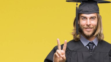 Genç yakışıklı adam uzun saçlı zafer işareti yapıyor parmak gösterilen kamera seyir gülümseyen izole arka plan üzerinde mezun oldu. İki numaralı.