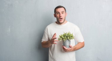 Bitki pot korkmuş şok bir sürpriz yüz, korkuyor ve heyecanlı korku ifadesi ile tutarak gri grunge duvarın üzerinden beyaz delikanlı