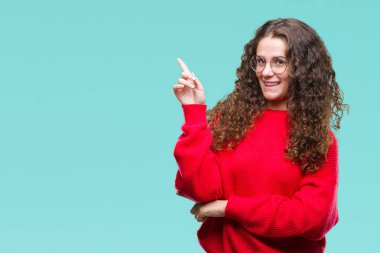 Kameraya bakarak yan el ve parmak ile işaret yüzünde kocaman bir gülümseme ile izole arka plan üzerinde gözlük ve kış kazak giyen güzel esmer kıvırcık saçlı Genç kız.