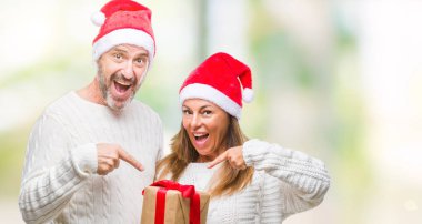 Orta yaş İspanyol çift giyen Noel şapka ve holding hediye izole arka el ve parmak ile işaret çok mutlu üzerinde