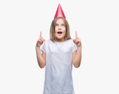 Genç güzel kız Doğum günü şapkası izole arka plan üzerinde hayret ve yukarıya bakıp parmakları ile işaret şaşırttı ve silah kaldırdı.