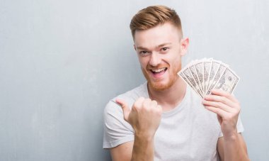 Genç Kızıl saçlı adam işaret ve başparmak kadar mutlu yüz gülümseyen tarafı ile gösterilen dolar tutan gri grunge duvarın üzerinden