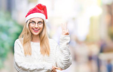 Mutlu yüz zafer işareti yaparken kameraya göz kırpıyor ile gülümseyen izole arka plan üzerinde Noel şapka giyen genç güzel sarışın kadın. İki numaralı.