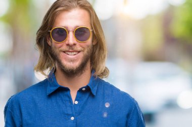 Genç yakışıklı adam uzun saçlı yüzünde mutlu ve güzel bir gülümseme ile izole arka plan üzerinde güneş gözlüğü takıyor. Şanslı kişi.