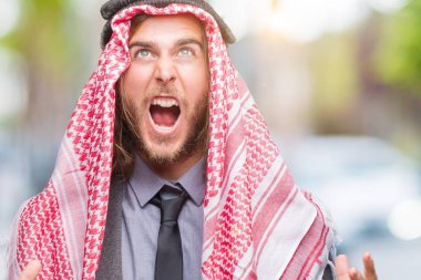 Genç yakışıklı Arap adam uzun saçlı izole arka plan çılgın ve kızgın bağırarak ve agresif ifade ve silah kaldırdı bağırmaya keffiyeh giyiyor. Hayal kırıklığı kavramı.
