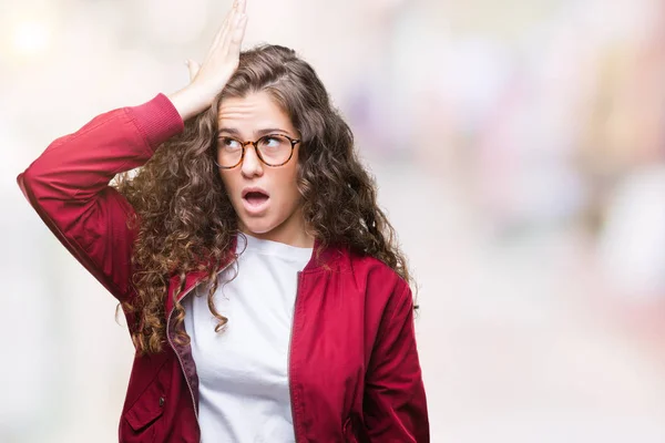 Beautiful brunette curly hair young girl wearing jacket and glasses over isolated background surprised with hand on head for mistake, remember error. Forgot, bad memory concept.