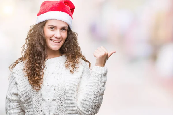 年轻的黑发女孩在孤立的背景上戴着圣诞帽微笑着 愉快的脸看着 用拇指指着一边 — 图库照片