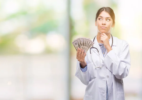 Young hispanic doctor woman holding dollars serious face thinking about question, very confused idea