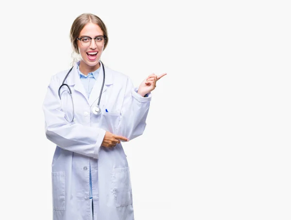 美しい金髪女医女性の顔に大きな笑みを浮かべて隔離された背景に医療制服を着てカメラを見て側の手と指で指す — ストック写真