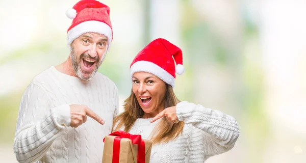 非常に幸せな手と指で指している孤立した背景の上クリスマス帽子 持株ギフトを着て中年ヒスパニック系カップル — ストック写真