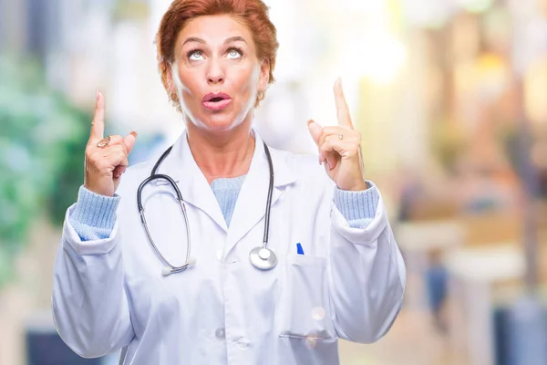 高级白种人医生妇女穿着医疗制服在孤立的背景惊讶和惊讶地抬起头来 用手指和举起的手臂指指点点 — 图库照片