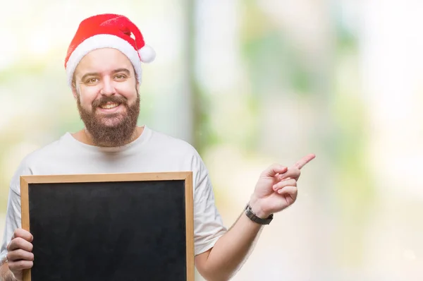 孤立した背景側の手と指で指す非常に幸せにクリスマス帽子持株黒板を着た若い白人ヒップ男 — ストック写真