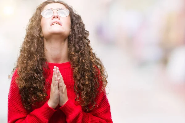 Beautiful brunette curly hair young girl wearing glasses and winter sweater over isolated background begging and praying with hands together with hope expression on face very emotional and worried. Asking for forgiveness. Religion concept.