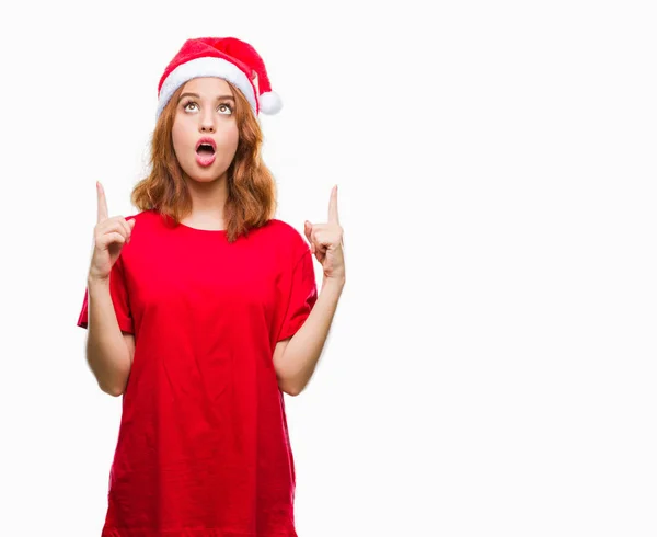 年轻美丽的女人在孤立的背景下戴着圣诞帽惊讶 惊讶地看着手指和举起的武器指向 — 图库照片