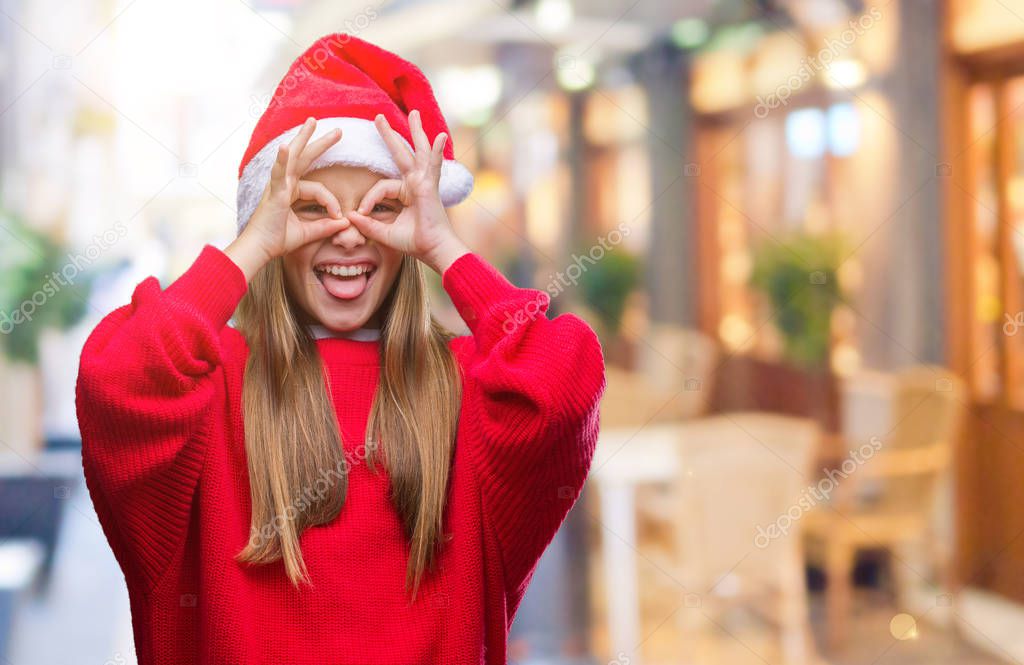 Young beautiful girl wearing christmas hat over isolated background doing ok gesture like binoculars sticking tongue out, eyes looking through fingers. Crazy expression.