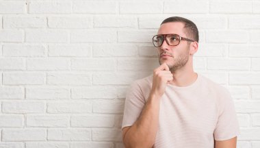 Soru, çok karışık fikir hakkında ciddi bir yüz düşünme genç beyaz adam ayakta beyaz tuğla duvar giyen üzerinde gözlük