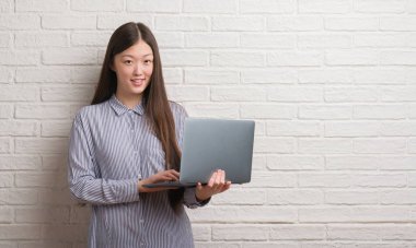 Genç Çinli kadın ayakta ve diş gösterilen kendine güvenen bir gülümsemeyle gülümseyerek mutlu bir yüz ile dizüstü bilgisayar kullanarak tuğla duvarın üzerinden