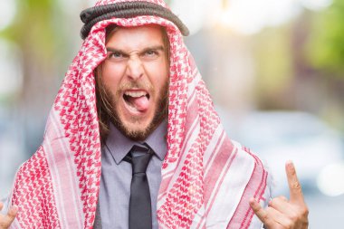 Genç yakışıklı Arap adam uzun saçlı rock simge elle yapıyor deli ifade ile bağırarak izole arka plan üzerinde keffiyeh giyiyor. Müzik yıldızı. Ağır kavramı.
