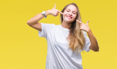 Kendinden emin gösterilen ve işaret parmakları diş ve ağız ile gülümseyen izole arka plan üzerinde rahat beyaz t-shirt giyen genç güzel sarışın bir kadın. Sağlık kavramı.