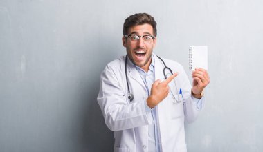 Yakışıklı genç doktor adam boş defter çok mutlu el ve parmak ile işaret tutan gri grunge duvarın üzerinden