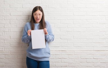 Boş kağıt bir sürpriz yüz, korkuyor ve heyecanlı korku ifadesi ile şok korkmuş tutan tuğla duvarın üzerinden Genç Çinli kadın