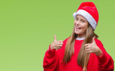 El ile pozitif jest yapıyor izole arka plan başarı işareti üzerinde Noel şapka giyen genç güzel kız gülümseyerek ve mutlu yaşasın. Neşeli bir ifadeyle, kazanan jest kameraya bakıyor.