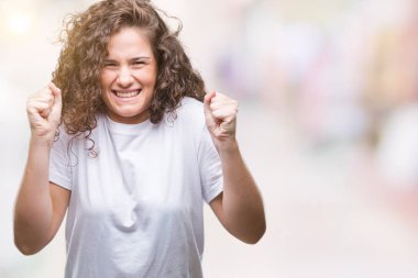 Güzel esmer kıvırcık saçlı Genç kız izole arka plan üzerinde rahat t-shirt giyen silah ile başarı için heyecanlı zafer kutluyor gülümseyen kaldırdı. Kazanan kavramı.