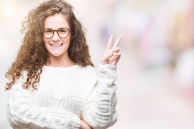 Güzel esmer kıvırcık saçlı Genç kız mutlu yüz zafer işareti yaparken kameraya göz kırpıyor ile gülümseyen izole arka plan üzerinde kış kazak giyiyor. İki numaralı.