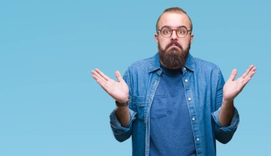 Üzerinde gözlük takan Genç beyaz hipster adam arka plan clueless ve şaşkın bir ifadeyle kollar ve eller kaldırdı izole. Şüphe kavramı.