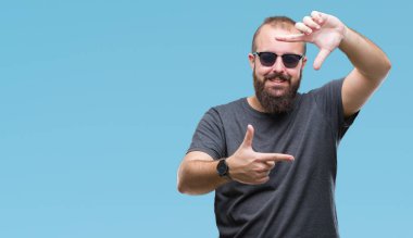Genç beyaz hipster adam elleriyle yapım çerçeve ve parmakları ile mutlu yüz gülümseyen izole arka plan üzerinde güneş gözlüğü takıyor. Yaratıcılık ve Fotoğrafçılık kavramı.