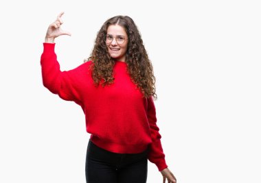 Güzel esmer kıvırcık saçlı Genç kız izole arka plan gülümseyen ve boyutu işaret parmaklı ararken ve kamera işi el ile kendinden emin eller üzerinde gözlük ve kış kazak giyiyor. Ölçü kavramı.