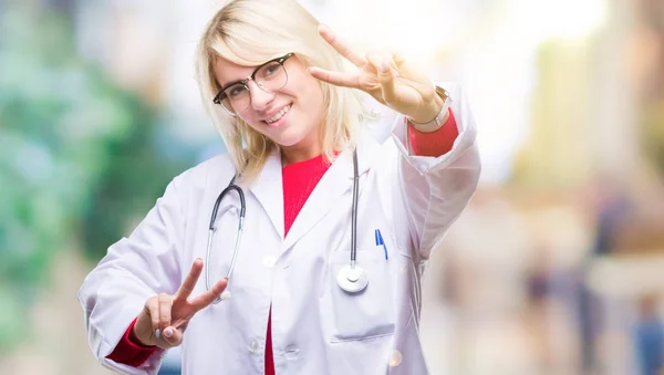 Young beautiful blonde doctor woman wearing medical uniform over isolated background smiling looking to the camera showing fingers doing victory sign. Number two.